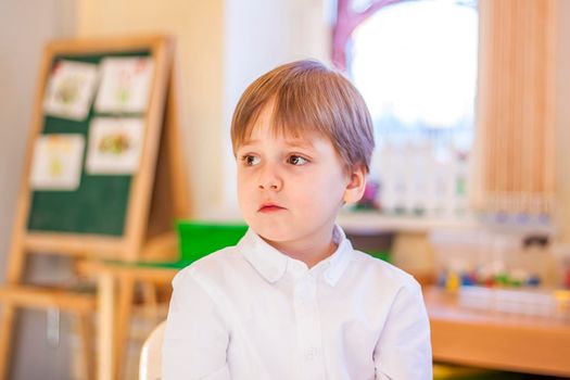 Elegantly dressed in a white shirt, a little boy is sitting in the classroom for lessons. portrait of a boy, blonde hair