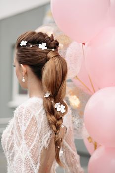 Back view of unrecognizable gorgeous brunette with beautiful hairdo and flowers in it posing in luxurious white dress with bunch on pink air balloons.