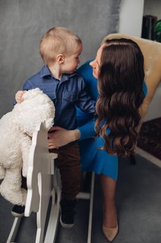 Full length studio portrait of gorgeous elegant brunette mom in denim dress with lovely laughing son on her hands. They smiling at camera.