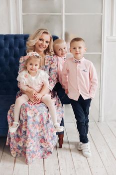 Lovely family sitting together, looking at camera in studio. Beautiful mother in dress hugging little daughter and two boys. Parent with children posing and smiling at home. Concept of love.