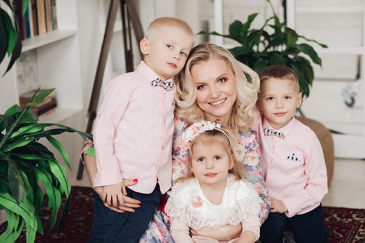 Front view of attractive mother posing with three cute kids at home. Young woman with sweet daughter and little boys looking at camera and smiling. Concept of childhood and motherhood.