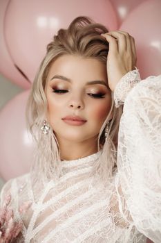 Fashion portrait of stunning blonde girl with hairstyle and professional make up in white transparent blouse holding bunch of pink colored flowers. Bunch of pink air balloons in background.