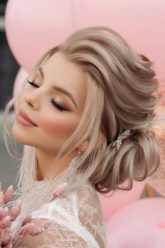 Fashion portrait of stunning blonde girl with hairstyle and professional make up in white transparent blouse holding bunch of pink colored flowers. Bunch of pink air balloons in background.