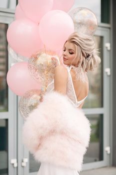 Back view of elegant attractive blonde woman with hairstyle and make up in white laced dress and luxurious pink fur coat posing in the street with bunch of air balloons.