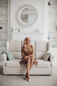 Gorgeous pretty lady sitting in luxury apartment and holding hands on knees. Beautiful young woman in elegant beige dress posing on white sofa. Happy blonde model relaxing after long working day.