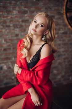 Gorgeous blonde woman sitting on high chair and thoughtfully looking at camera. Young pretty lady in red dress with naked shoulder touching floor by feet. Elegant model holding hand near face posing.