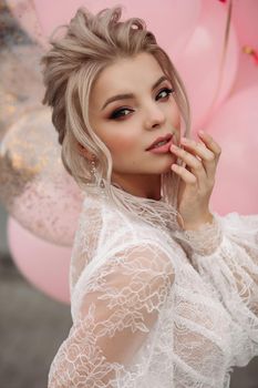 Fashion portrait of stunning blonde girl with hairstyle and professional make up in white transparent blouse holding bunch of pink colored flowers. Bunch of pink air balloons in background.