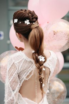 Back view of unrecognizable gorgeous brunette with beautiful hairdo and flowers in it posing in luxurious white dress with bunch on pink air balloons.