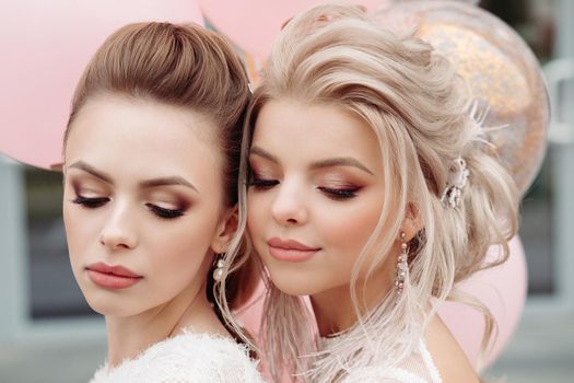 Portrait of attractive, stunning ladies with professional make up and hairstyles wearing white laced outlooks. Brunette and blonde looking down showing their make up. Pink air balloons in background.