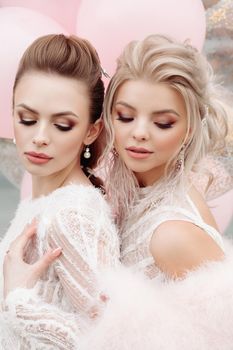 Portrait of attractive, stunning ladies with professional make up and hairstyles wearing white laced outlooks. Brunette and blonde looking down showing their make up. Pink air balloons in background.