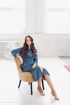 Full length of attractive elegant brunette young woman in stylish casual denim dress and beige heels reading a fashion magazine. She is in living room next to the bookshelf and plants.