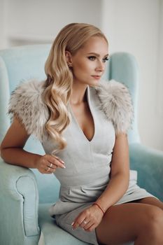 Front view of beautiful blonde in blue short dress and heels sitting on dressing table and posing. Attractive woman with long legs looking down and smiling at home. Concept of beauty and glamour.