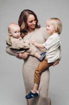 Young beautiful mother wearing long dress keeping two children and posing on grey isolated background. Pretty woman hugging, looking at each other and laughing in studio. Concept of motherhood.