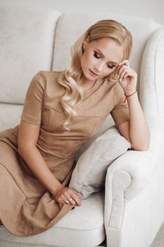 Gorgeous pretty lady sitting in luxury apartment and holding hands on knees. Beautiful young woman in elegant beige dress posing on white sofa. Happy blonde model relaxing after long working day.