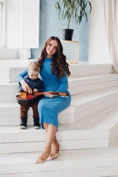 Full length portrait of gorgeous elegant brunette woman in blue dress and beige heels playing musical instrument with her little cute son. They sit on steps in the living room. Mother is smiling at camera.