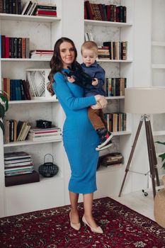 Stunning young slim brunette mother in casual blue dress and heels sitting on the floor with her adorable son playing with wood horse.
