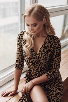 View from side of attractive calm lady wearing leopard dress sitting on windowsill and posing. Beautiful female model looking aside and waiting. Concept of beauty and elegance.