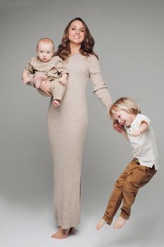 Happy young mother posing at camera together with her kids. Beautiful parent in casual clothes holding little baby and giving hand to other one. Brunette woman smiling and standing with cute sons.