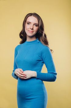 Studio portrait of attractive brunette woman with wavy hair in casual blue dress with hand over hand on her tummy. She is posing over plain yellow background. Isolate. Smiling calmly at camera.