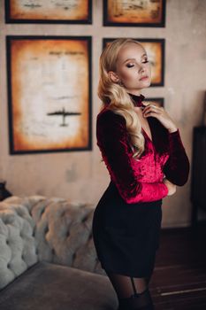 Close up from above of beautiful young woman in dark red dress looking at camera. Happy blonde girl with long hair with make up smiling. Portrait of gorgeous model sitting and posing in elegant look.