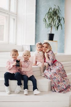 Gorgeous blonde mother in dress hugging with lovely daughter while two sons in similar clothes playing with film camera sitting on steps at home. Happy family concept.