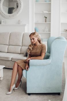 Gorgeous pretty lady sitting in luxury apartment and holding hands on knees. Beautiful young woman in elegant beige dress posing on white sofa. Happy blonde model relaxing after long working day.
