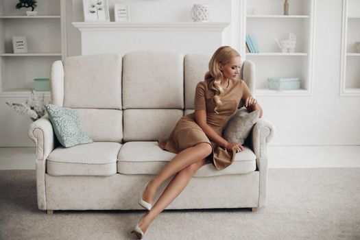 Gorgeous pretty lady sitting in luxury apartment and holding hands on knees. Beautiful young woman in elegant beige dress posing on white sofa. Happy blonde model relaxing after long working day.