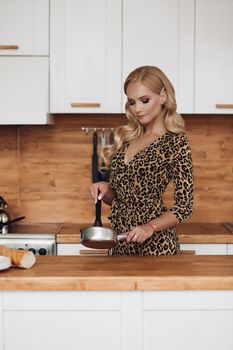Stock photo portrait of stunning elegant blonde woman with hairstyle wearing leopard printed dress cooking in the kitchen at home. She looks gorgeous and preparing breakfast in the kitchen.