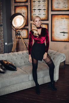 Full length portrait of stunning blonde woman with make up and hairstyle posing in elegant sexy dress of red and black color. She is wearing sexy black tights and boots. Posing beside the modern grey couch.