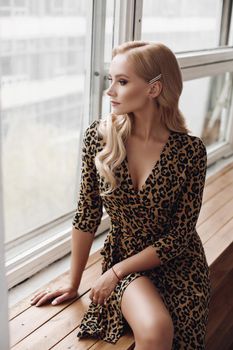 View from side of attractive calm lady wearing leopard dress sitting on windowsill and posing. Beautiful female model looking aside and waiting. Concept of beauty and elegance.