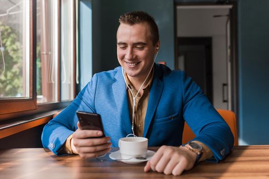A stylish happy guy businessman of European appearance in a jacket and shirt sits at a table in a cafe on a coffee break, holds a smartphone or mobile phone in his hand.