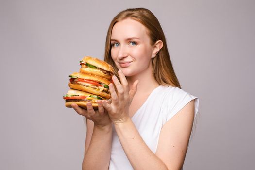 Selective focus of big tasty burgers in hands of amazed girl on grey isolated background. Shocked woman with big eyes looking at camera and eating delicious fast food. Concept of size and food.