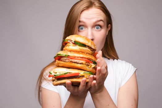 Selective focus of big tasty burgers in hands of amazed girl on grey isolated background. Shocked woman with big eyes looking at camera and eating delicious fast food. Concept of size and food.