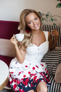 Stunning young blonde woman in beautiful summer dress and heels drinking tea or coffee sitting on window sill in a cafe beside table with cupcakes. She is looking through the window on sunny street.