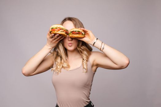 Selective focus of big tasty burgers in hands of amazed girl on grey isolated background. Shocked woman with big eyes looking at camera and eating delicious fast food. Concept of size and food.