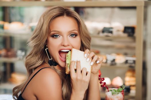 Close up of pretty young girl holding tasty sandwich in her hands. Beautiful woman sitting in restaurant and enjoying lunch. Adorable blonde lady with manicure eating unhealthy but delicious food.