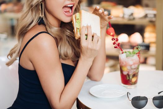 Medium close-up portrait of beautiful young fashion woman biting fresh appetizing sandwich. Portrait of smiling adorable female posing enjoying eating fast food in cafe