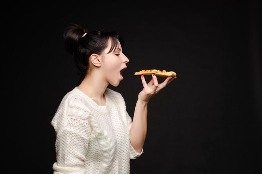 Side view portrait of brunette young girl in white sweater with tail biting delicious piece of Italian pizza. Isolate on black. Fast food concept.