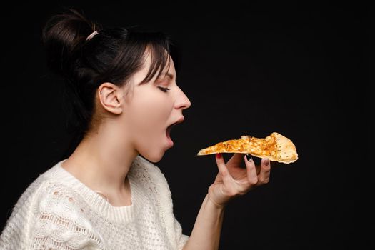 Close-up face of hungry young woman open mouth ready biting appetizing piece of pizza. Casual attractive young female enjoying eating fast food isolated at studio black background side view