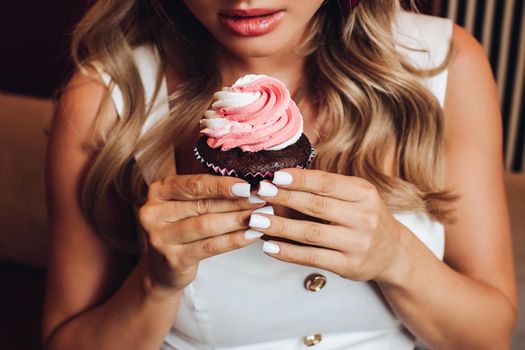 View from above of pretty girl keeping delicious pink cupcake in hands. Young female looking at tasty sweet chocolate cake and enjoying smell. Concept of pastry and desserts.