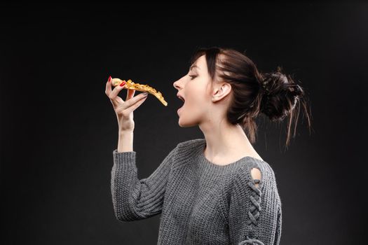 Side view portrait of brunette young girl in white sweater with tail biting delicious piece of Italian pizza. Isolate on black. Fast food concept.