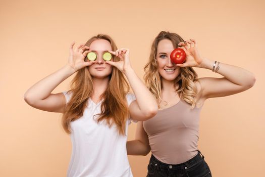 Front view of two healthy women keeping cucumber and apple and closing eyes with fresh vegetables and fruits on isolated background. Girls eating vitamins and keeping diet and fit. Concept of energy.