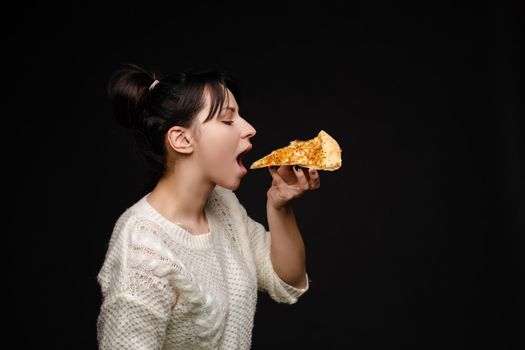 Side view portrait of brunette young girl in white sweater with tail biting delicious piece of Italian pizza. Isolate on black. Fast food concept.