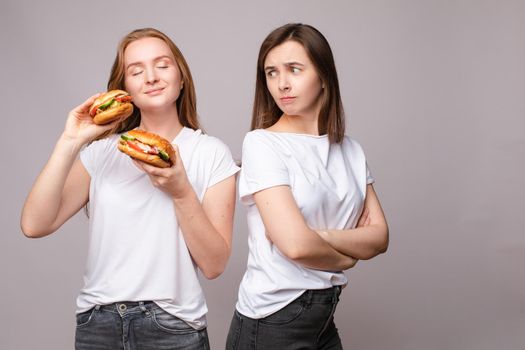 Two frowning young beautiful woman holding appetizing harmful burger looking at camera. Disapproving European female posing with unhealthy fast food isolated at gray studio background medium shot