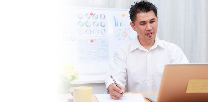 Asian businessman talking with team via video conference writing note on paper. business people using laptop  for online meeting. Business work from home concept.