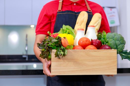 Grocery delivery courier man in red uniform with grocery box with fresh fruit and vegetable. Deliver man in red uniform and apron handling wooden crate of food, fruit, vegetable with kitchen background. Delivery Concept