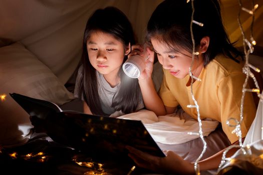 Family concept. elder sister and sister reading book with flashlight together in children tent before bedtime. Happy elder sister read story book to her sister in bed sheet tent with 