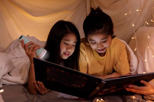 Family concept. elder sister and sister reading book with flashlight together in children tent before bedtime. Happy elder sister read story book to her sister in bed sheet tent
