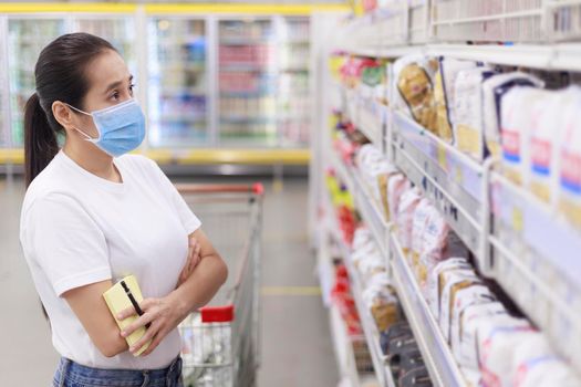 Asian woman wear face mask push shopping cart in supermarket. female looking grocery to buy something During coronavirus crisis or covid19 outbreak. Women wearing protective face mask new normal concept