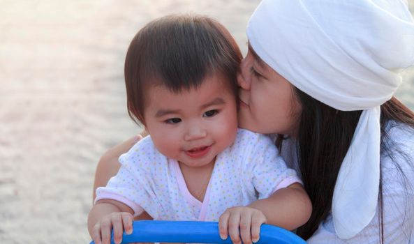 Close up portrait beautiful young asian mother kissing her little baby in park. Happy lovely mother kiss her little daughter. Smiling little girl play at playground with her mother.